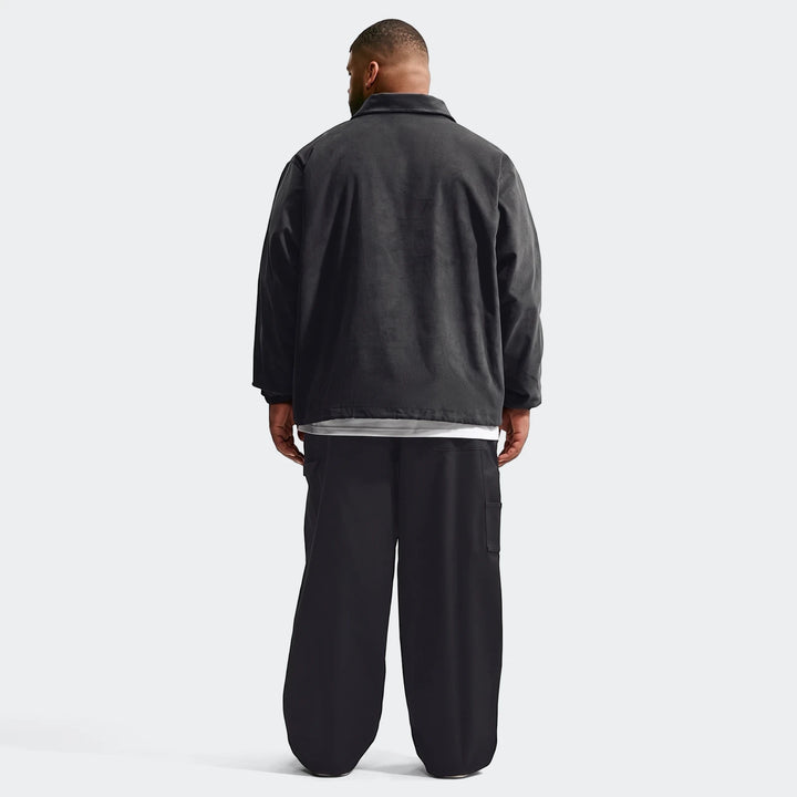 Back view of a man wearing a black Nike Club Men's Corduroy Coaches Jacket and black wide-leg cargo pants on a white