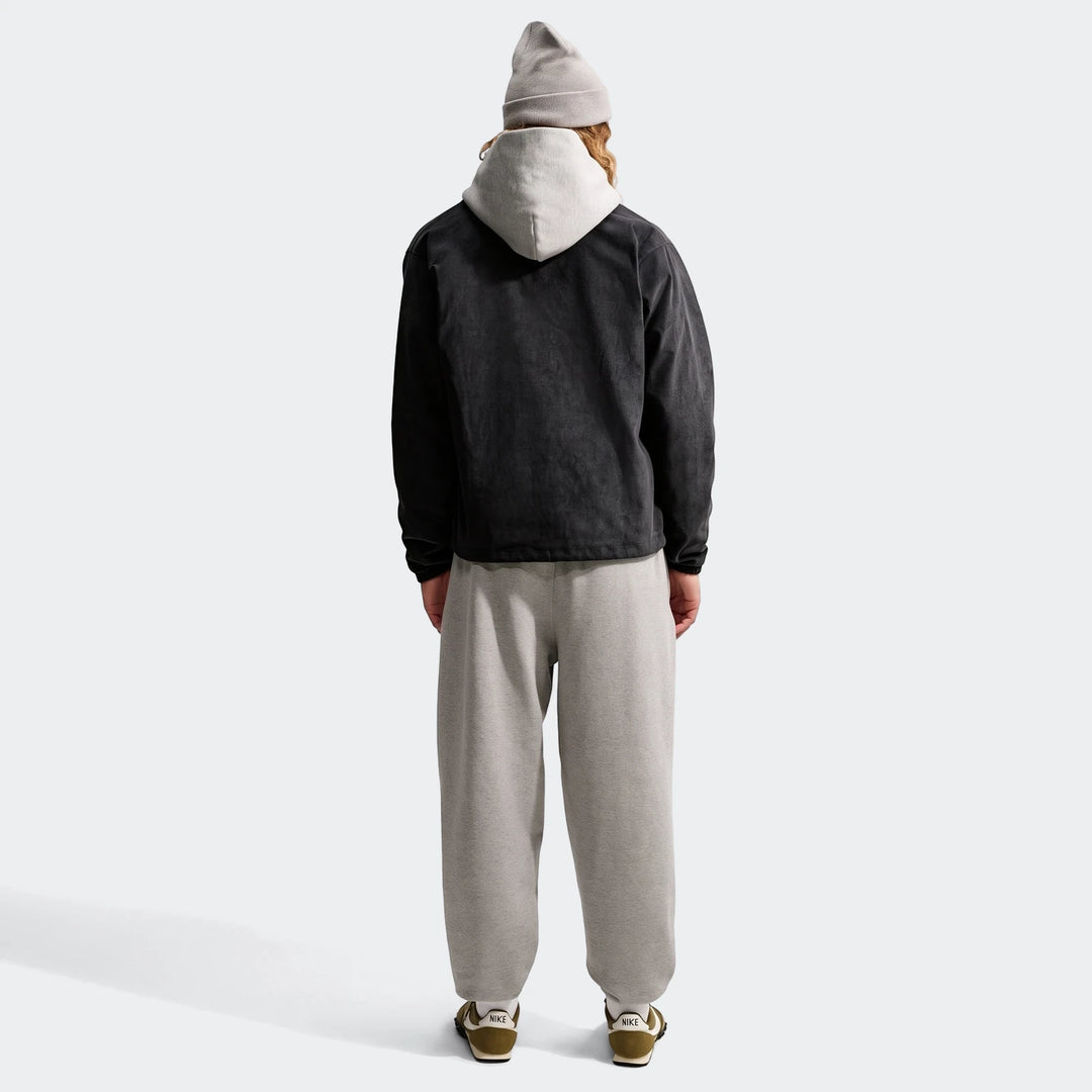 Back view of a man wearing a black Nike Club men's corduroy coaches jacket, a grey hoodie, grey sweatpants, and green Nike