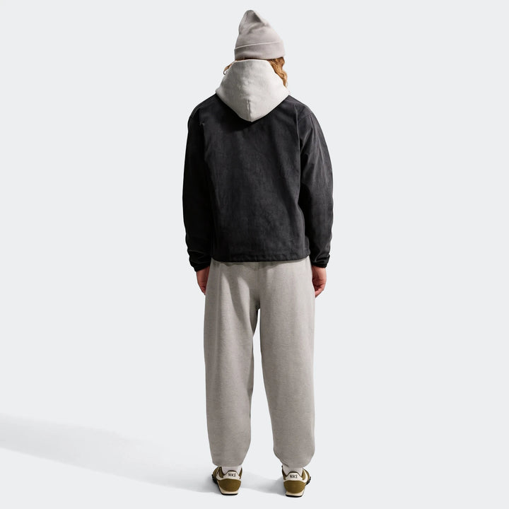 Back view of a man wearing a black Nike Club men's corduroy coaches jacket, a grey hoodie, grey sweatpants, and green Nike