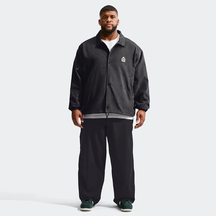 Full-body shot of a man wearing a black Nike Club men's corduroy coaches jacket, black wide-leg pants, and green sneakers.