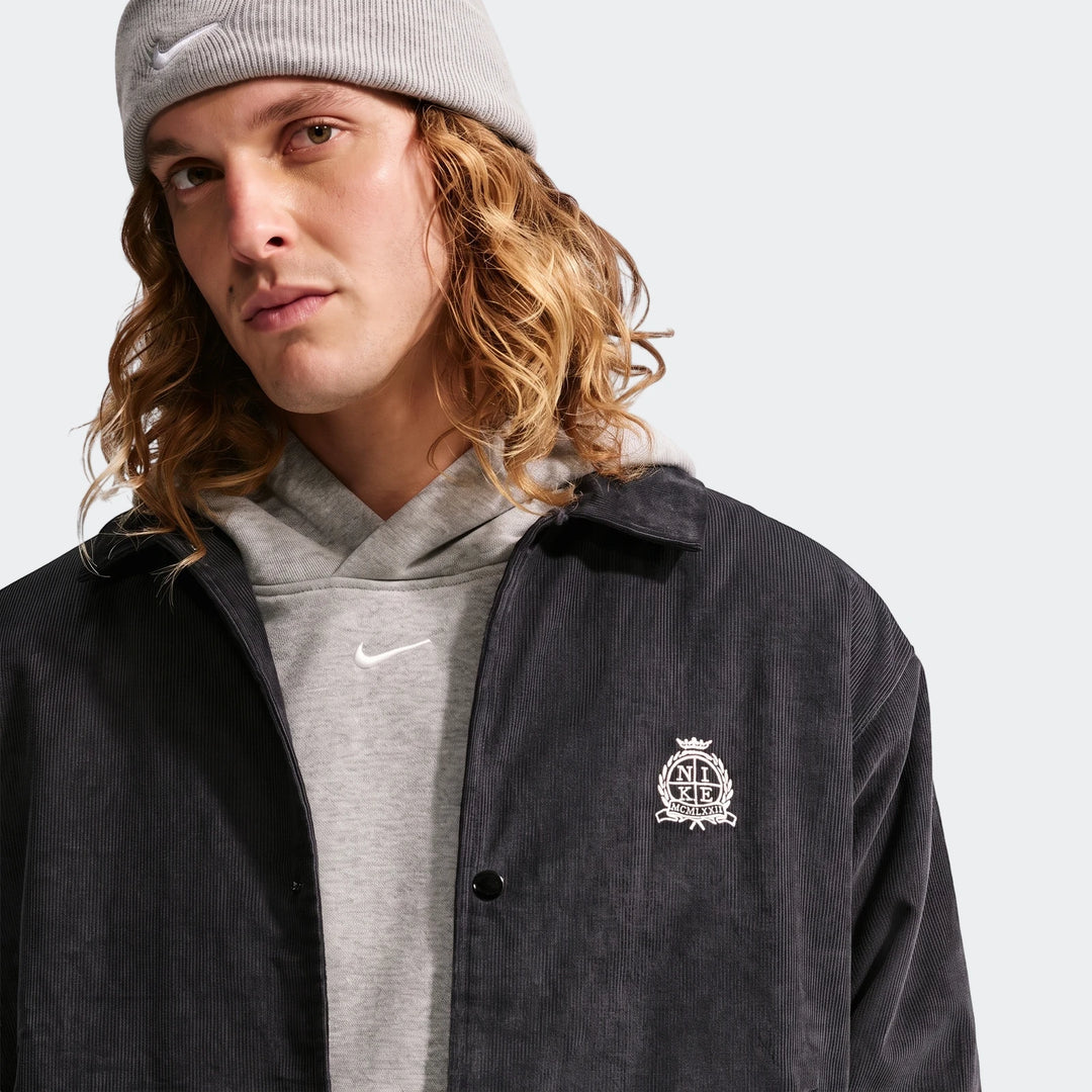 A man wears a black corduroy Nike Club Men's Coaches Jacket over a gray hoodie and beanie, looking directly forward.
