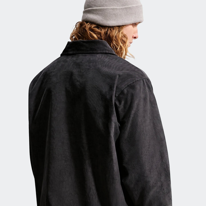 Men's Nike Club corduroy coaches jacket in dark grey, worn by a model with a grey beanie, viewed from the back-right.