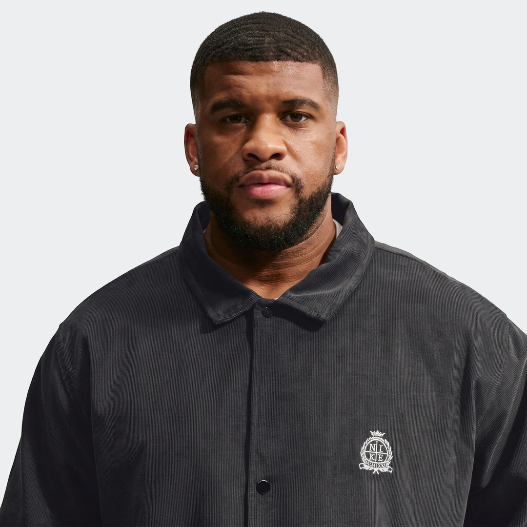 Front view of a man wearing a dark grey Nike Club Men's Corduroy Coaches Jacket with an embroidered logo on the chest.