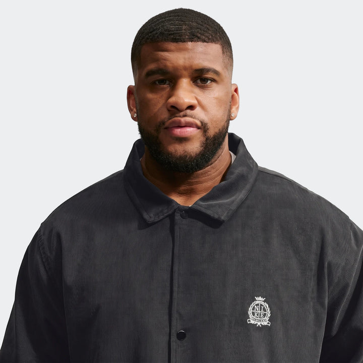 Front view of a man wearing a dark grey Nike Club Men's Corduroy Coaches Jacket with an embroidered logo on the chest.