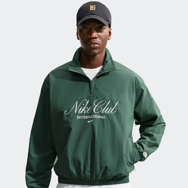 Man wearing a dark green Nike Club Men's 1/2 Zip Jacket with a white 'Nike Club INTERNATIONAL' logo and a black cap.