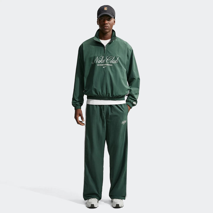 A male model showcases the Nike Club Men's 1/2 Zip Jacket in green, paired with matching green tracksuit pants, standing