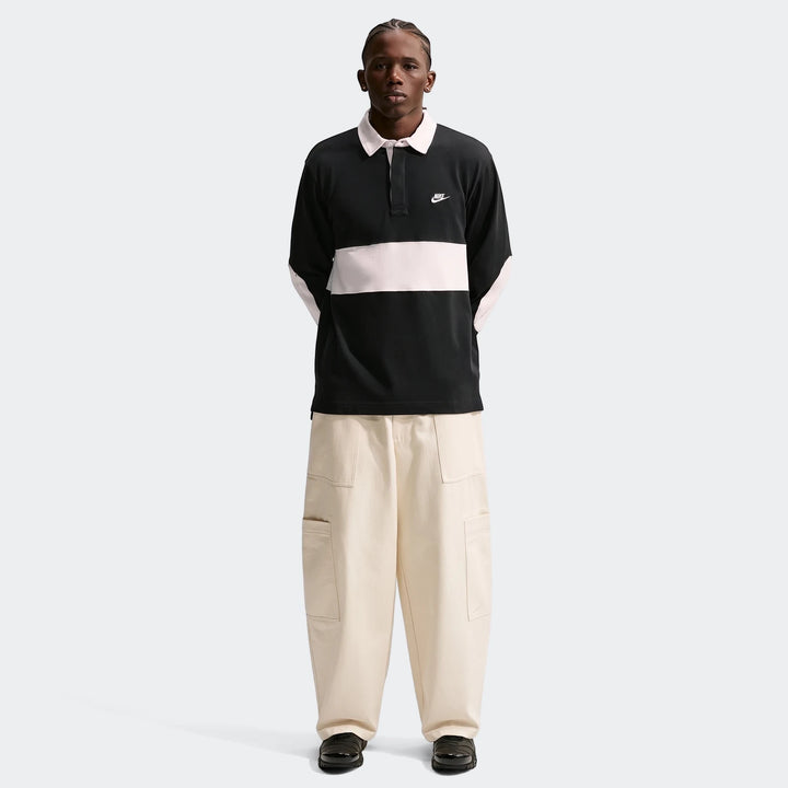 A full-body image of a man wearing a black Nike Club Men's Long-Sleeve Rugby Top with a light pink stripe, cream cargo