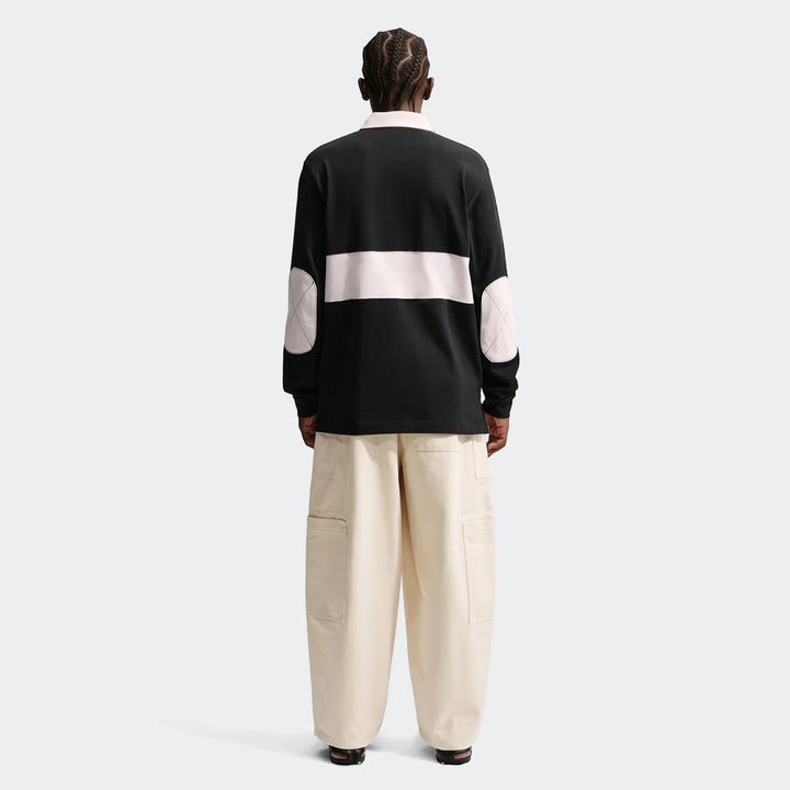 Back view of a model wearing a black Nike Club Men's Long-Sleeve Rugby Top with a light pink stripe and elbow patches,