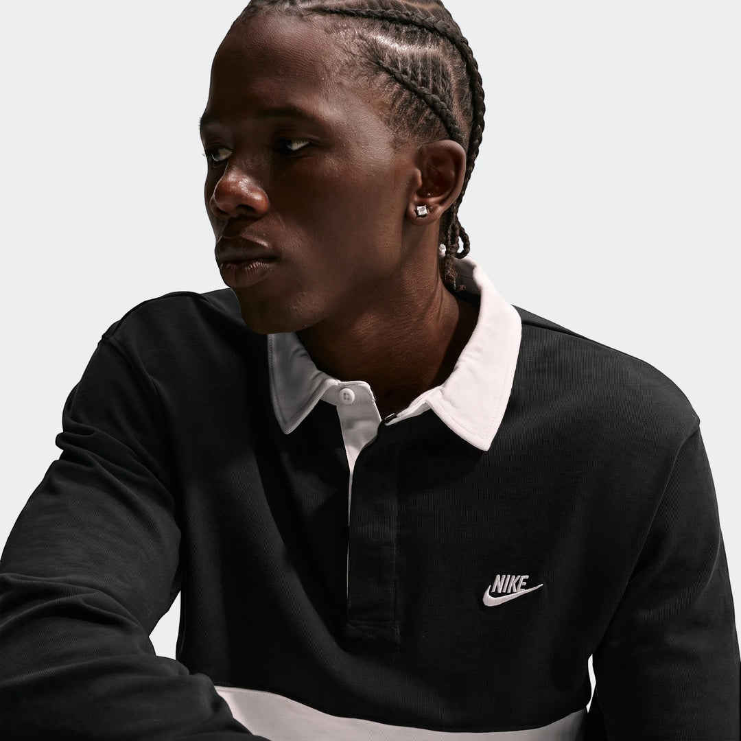 A man wears a Nike Club long-sleeve rugby top in black with a white collar and stripe, featuring an embroidered logo.