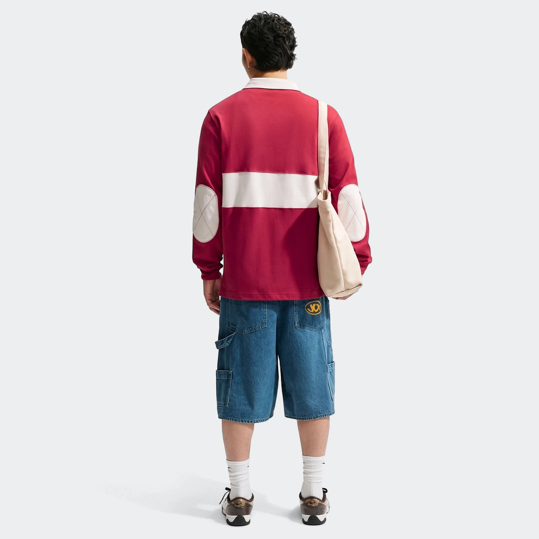 Rear view of a man wearing a red Nike Club long-sleeve rugby top with white stripe, blue denim shorts, and sneakers.