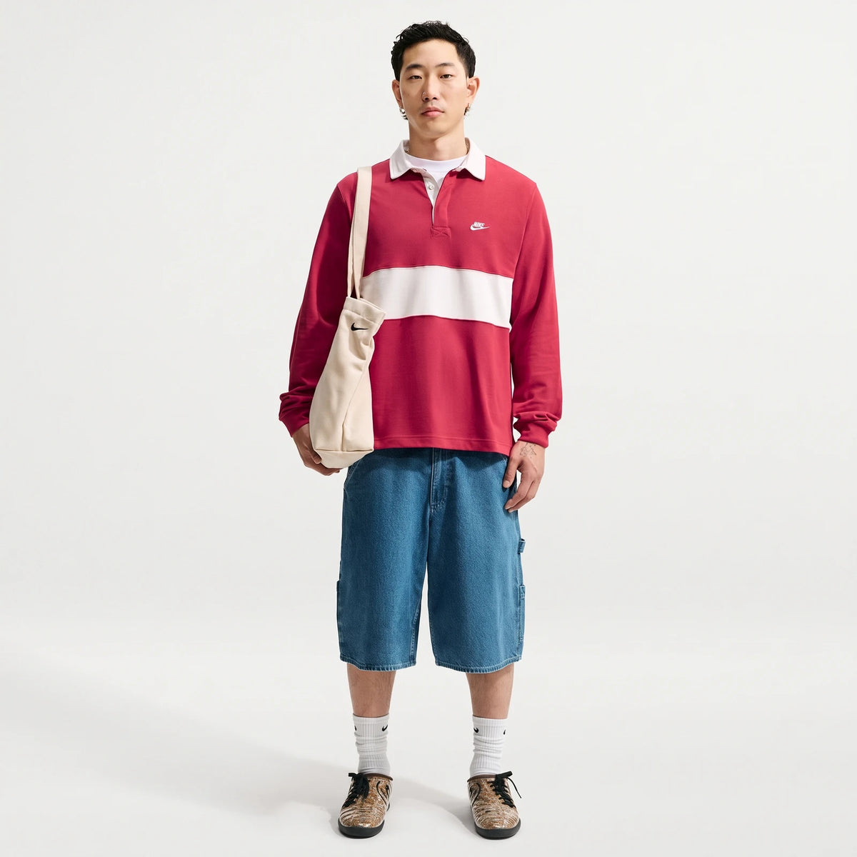 Male model wearing a red and white striped Nike Club Men's Long-Sleeve Rugby Top, blue denim shorts, and holding a beige