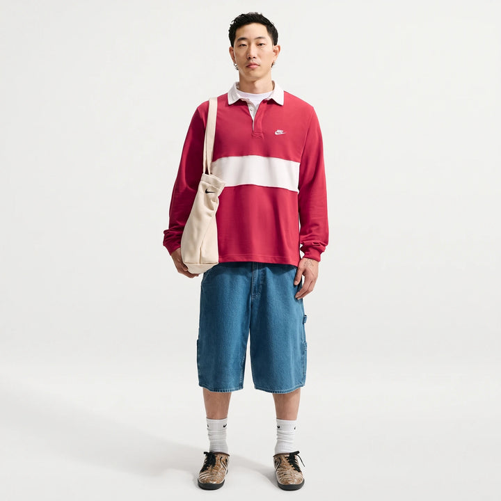 Male model wearing a red and white striped Nike Club Men's Long-Sleeve Rugby Top, blue denim shorts, and holding a beige