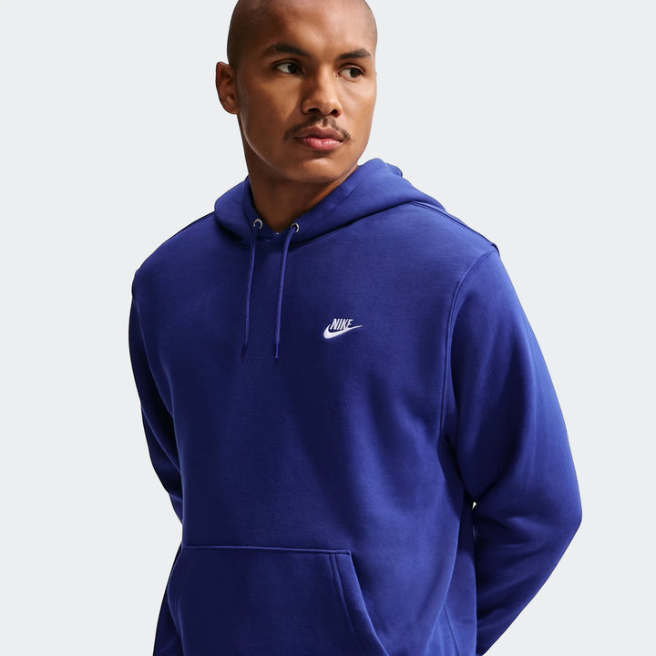 Man wearing a blue Nike Club Pullover Fleece Hoodie, posing from the waist up on a neutral background.