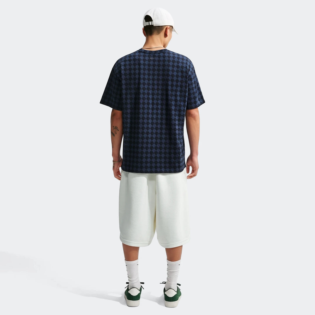 Rear view of a male model wearing a dark blue and black houndstooth Nike Club Rules T-shirt, white shorts, white cap, and