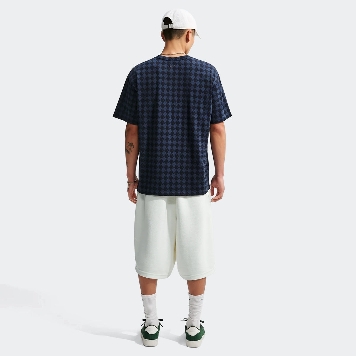 Rear view of a male model wearing a dark blue and black houndstooth Nike Club Rules T-shirt, white shorts, white cap, and
