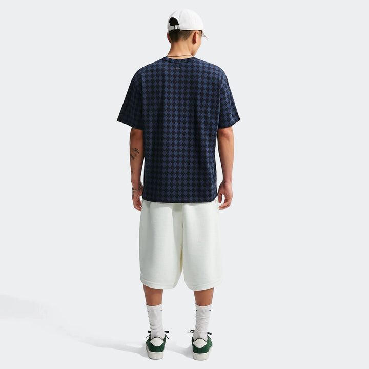 Rear view of a male model wearing a dark blue and black houndstooth Nike Club Rules T-shirt, white shorts, white cap, and