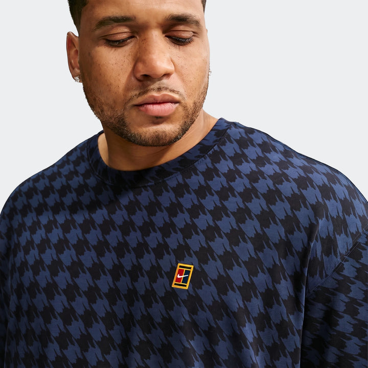 A close-up of a man wearing a blue and black Nike Club Rules T-Shirt with a houndstooth pattern and an embroidered logo.