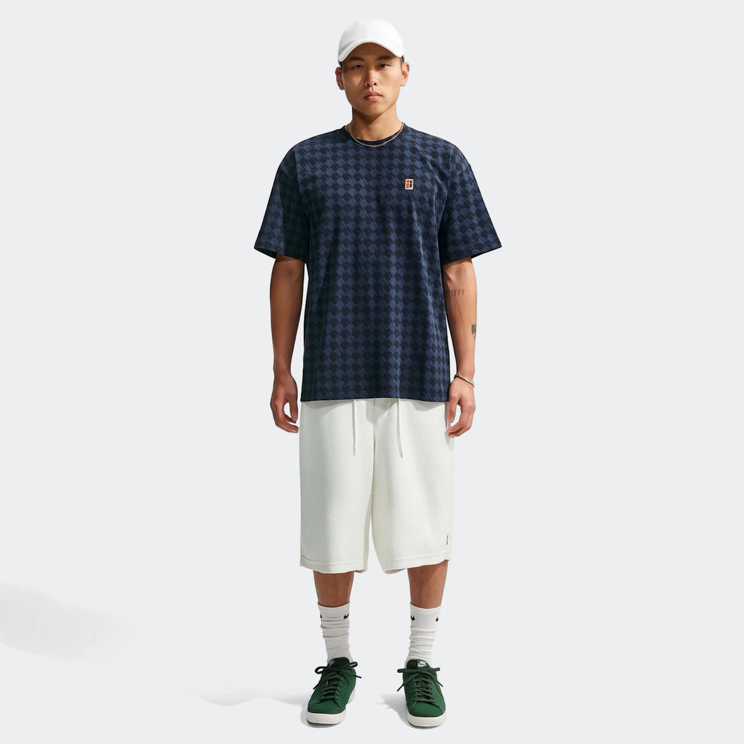 Young man wearing a blue and black houndstooth Nike Club Rules T-Shirt, white shorts, and green sneakers against a plain