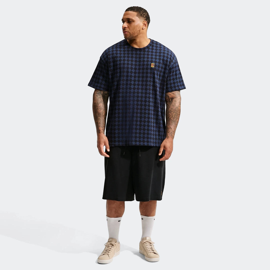 A male model wears a navy and black houndstooth Nike Club Rules T-Shirt, black shorts, and beige sneakers, standing
