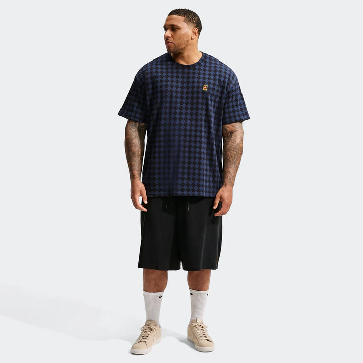 A male model wears a navy and black houndstooth Nike Club Rules T-Shirt, black shorts, and beige sneakers, standing