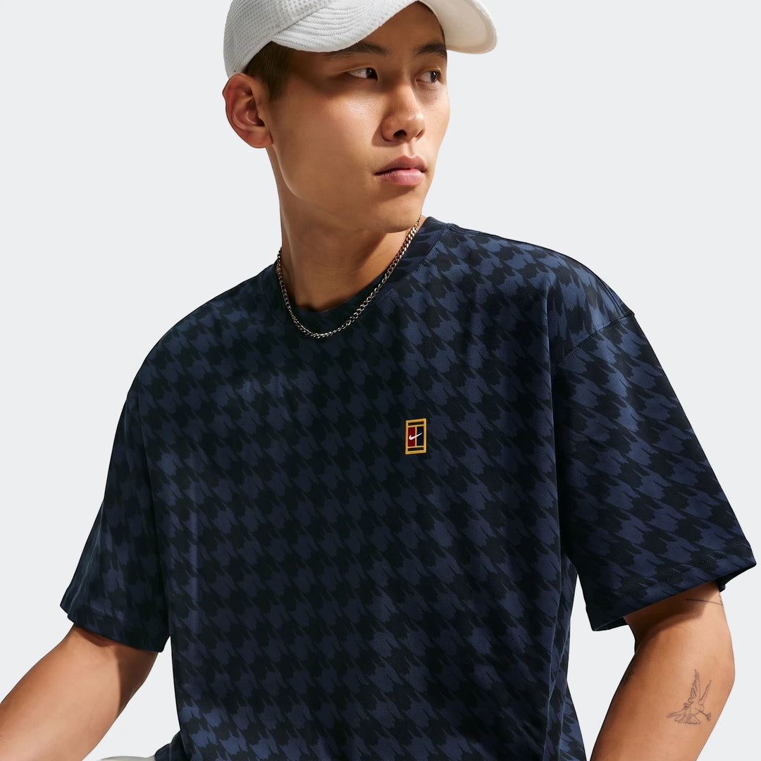 Young man wears a navy blue Nike Club Rules T-shirt with a houndstooth pattern and iconic tennis court logo.