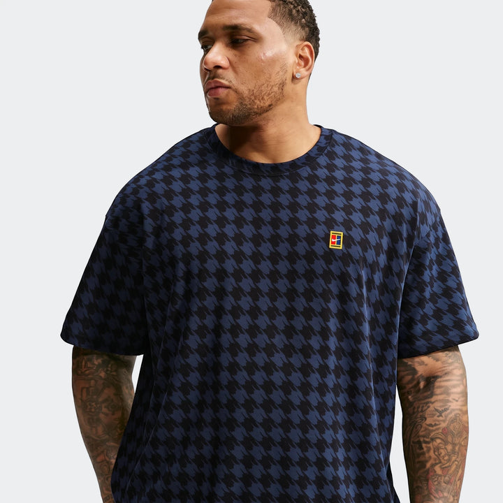 A man wears a black and blue houndstooth Nike Club Rules T-Shirt, featuring the iconic tennis court logo on the chest. The