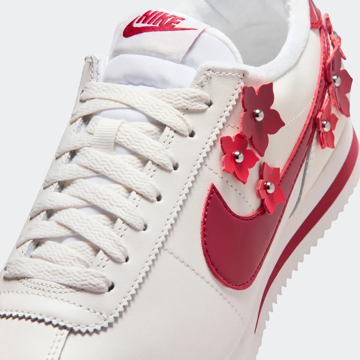 Close-up of a white Nike Cortez Leather Women's Shoe with a red Nike swoosh and red floral appliqués with silver studs.