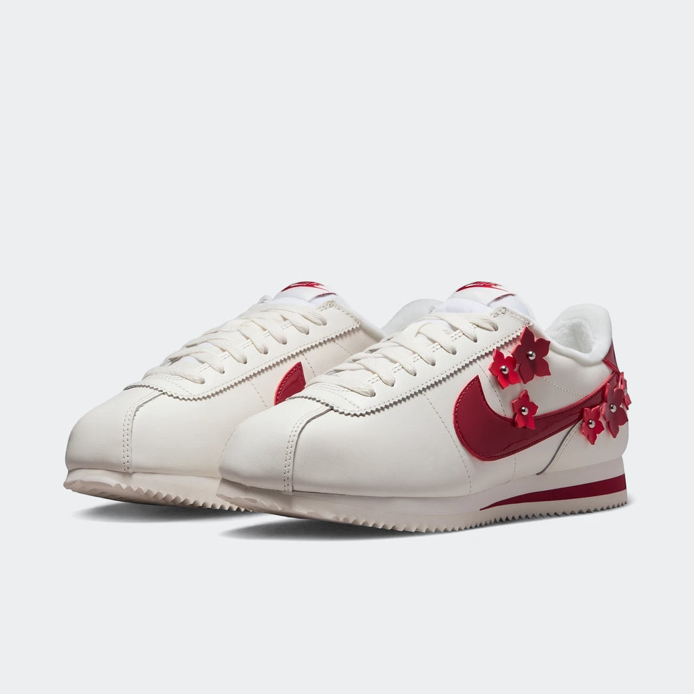 Pair of Nike Cortez Leather Women's Shoes in cream with red swooshes and delicate flower embellishments, displayed from a