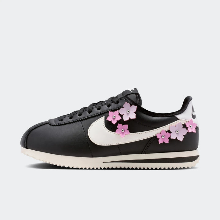 Black leather Nike Cortez Women's Shoes with white swoosh and pink cherry blossom flower embellishments, shown in a side