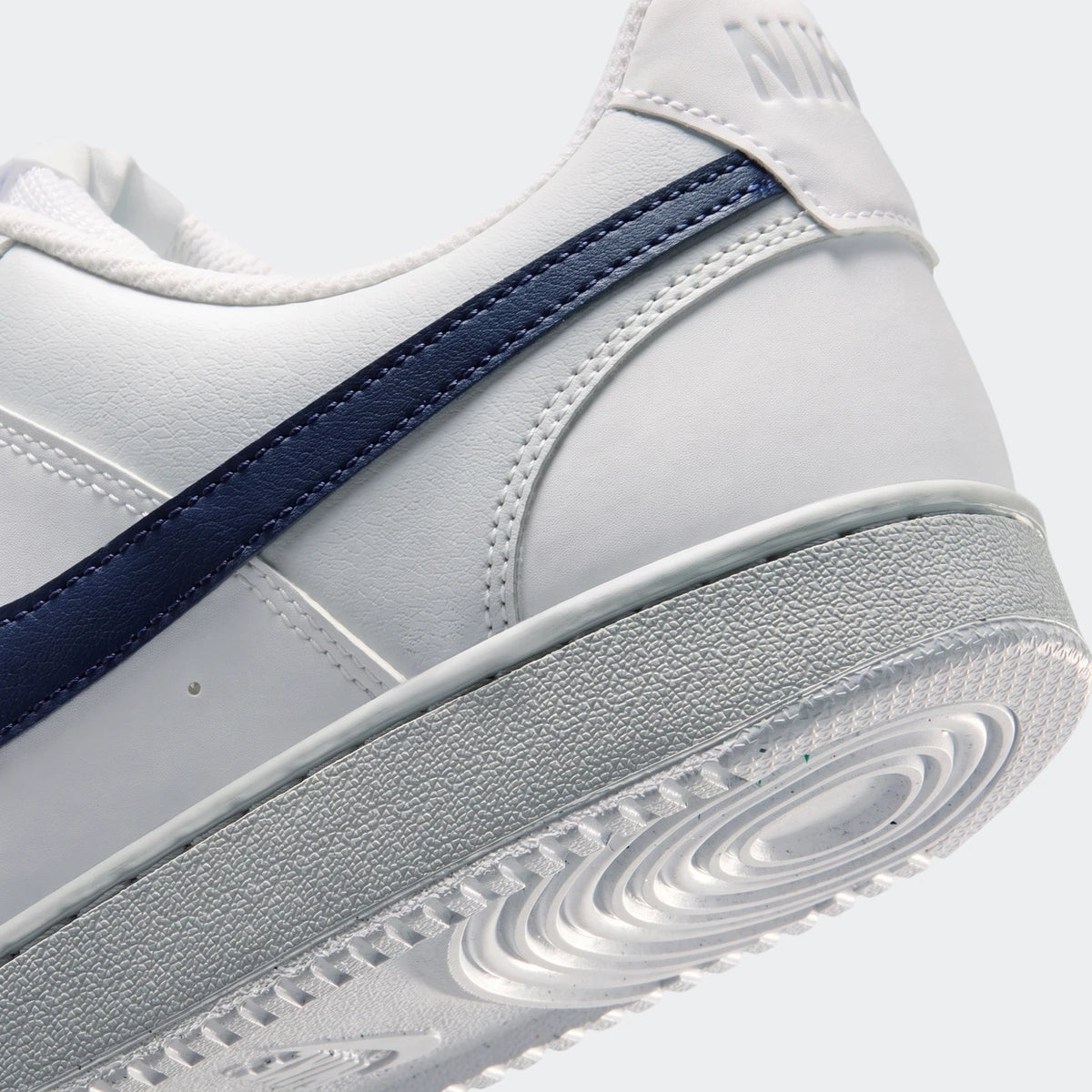 White Nike Court Vision Low men's shoes with a navy blue swoosh stripe, viewed from the rear-side, highlighting the heel and