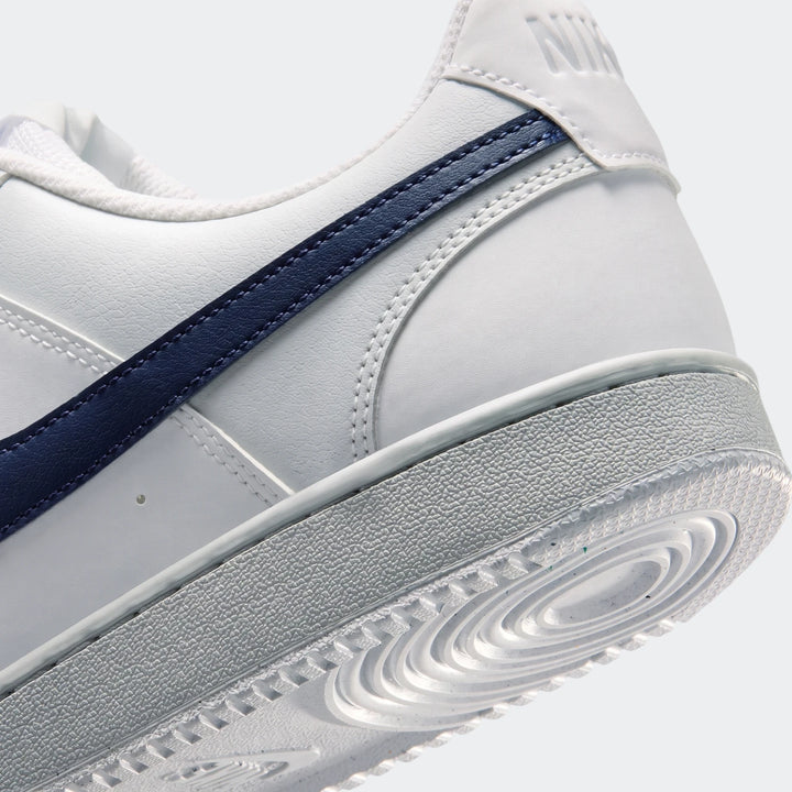White Nike Court Vision Low men's shoes with a navy blue swoosh stripe, viewed from the rear-side, highlighting the heel and