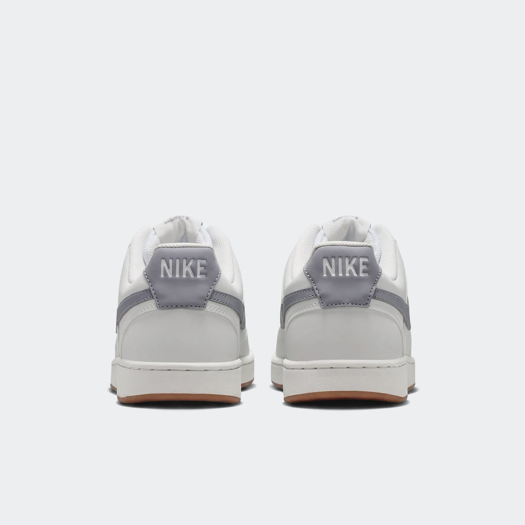 Rear view of two white Nike Court Vision Low men's shoes with grey heel accents and brown rubber soles, on a white