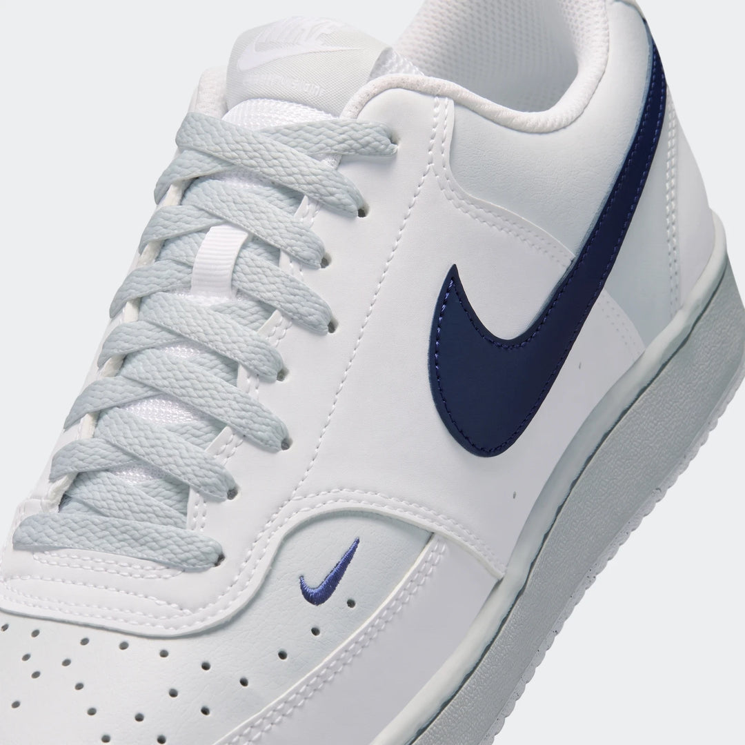 Nike Court Vision Low men's shoes in white with navy swoosh and light blue laces, shown from a front-side angle.