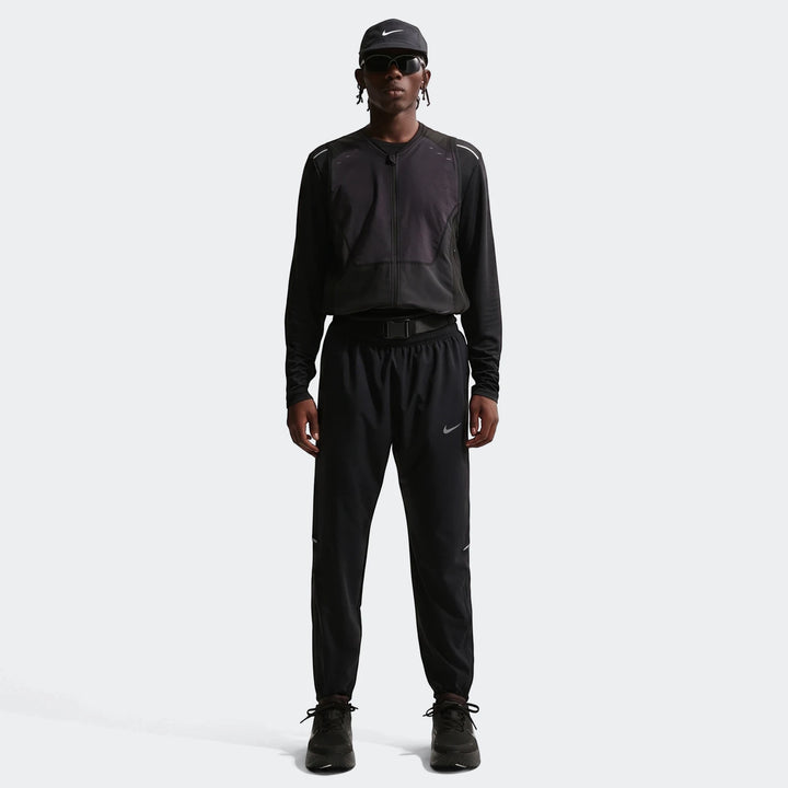 Full-body view of a male model wearing black Nike Df Miler Woven Pants, a black long-sleeve top, vest, cap, and sunglasses.