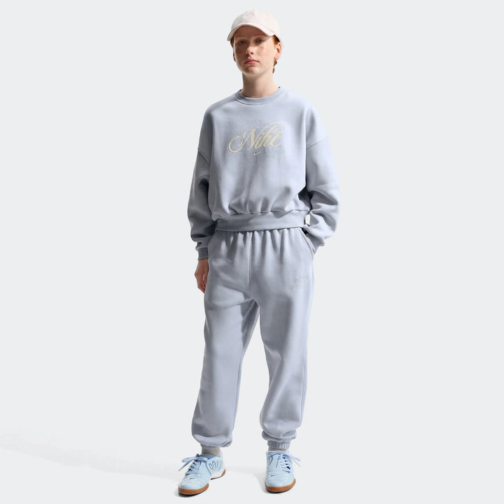 Full body shot of a female model wearing a light blue Nike fleece sweatshirt, matching sweatpants, light blue sneakers, and
