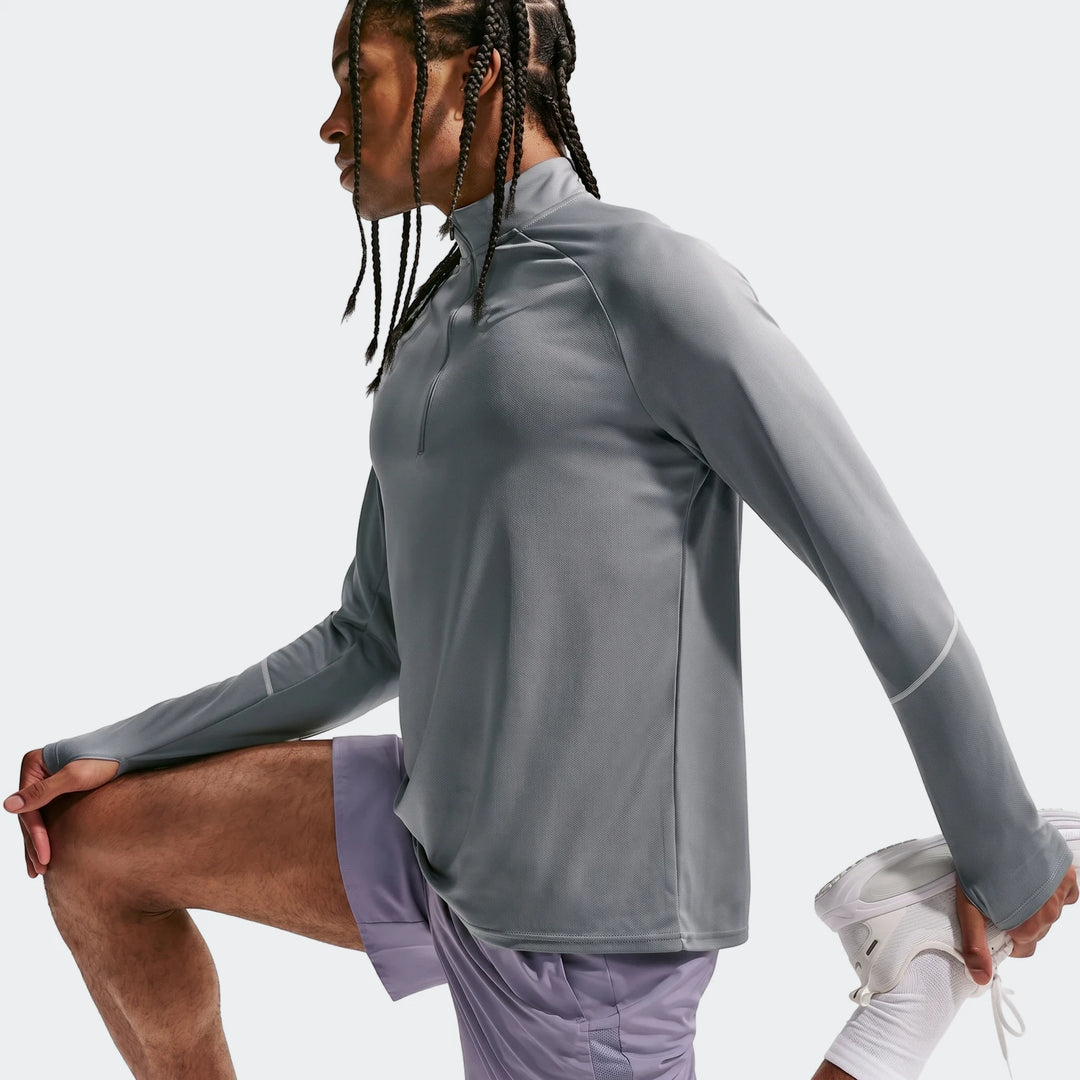 Man wearing a grey Nike Miler Dri-Fit 1/2-Zip Running Top with thumbholes and purple shorts, stretching in a studio.