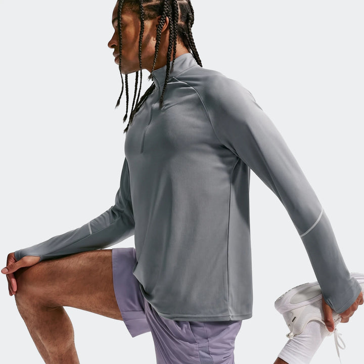 Man wearing a grey Nike Miler Dri-Fit 1/2-Zip Running Top with thumbholes and purple shorts, stretching in a studio.