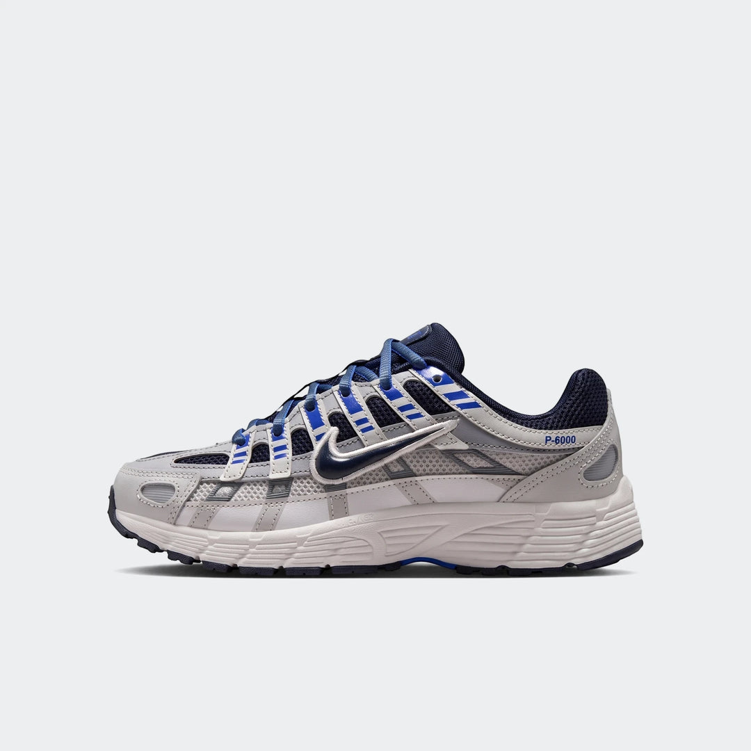 Side view of a Nike P-6000 Big Kids' Shoe in white, gray, and blue, featuring mesh and leather overlays with visible lacing.