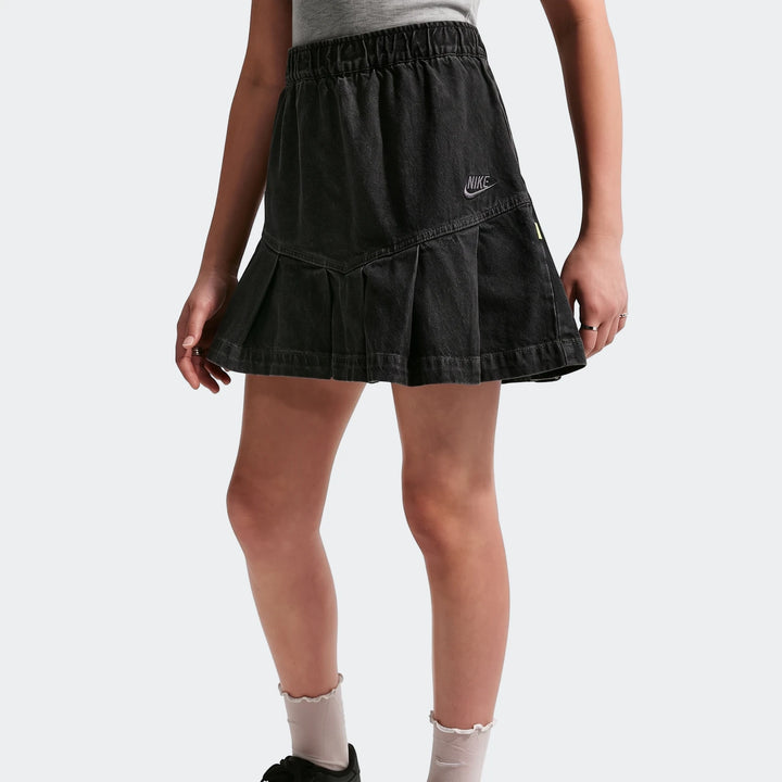 Black Nike Sportswear Big Kids' denim pleated skirt, worn by a child. Features an elastic waistband and embroidered logo.