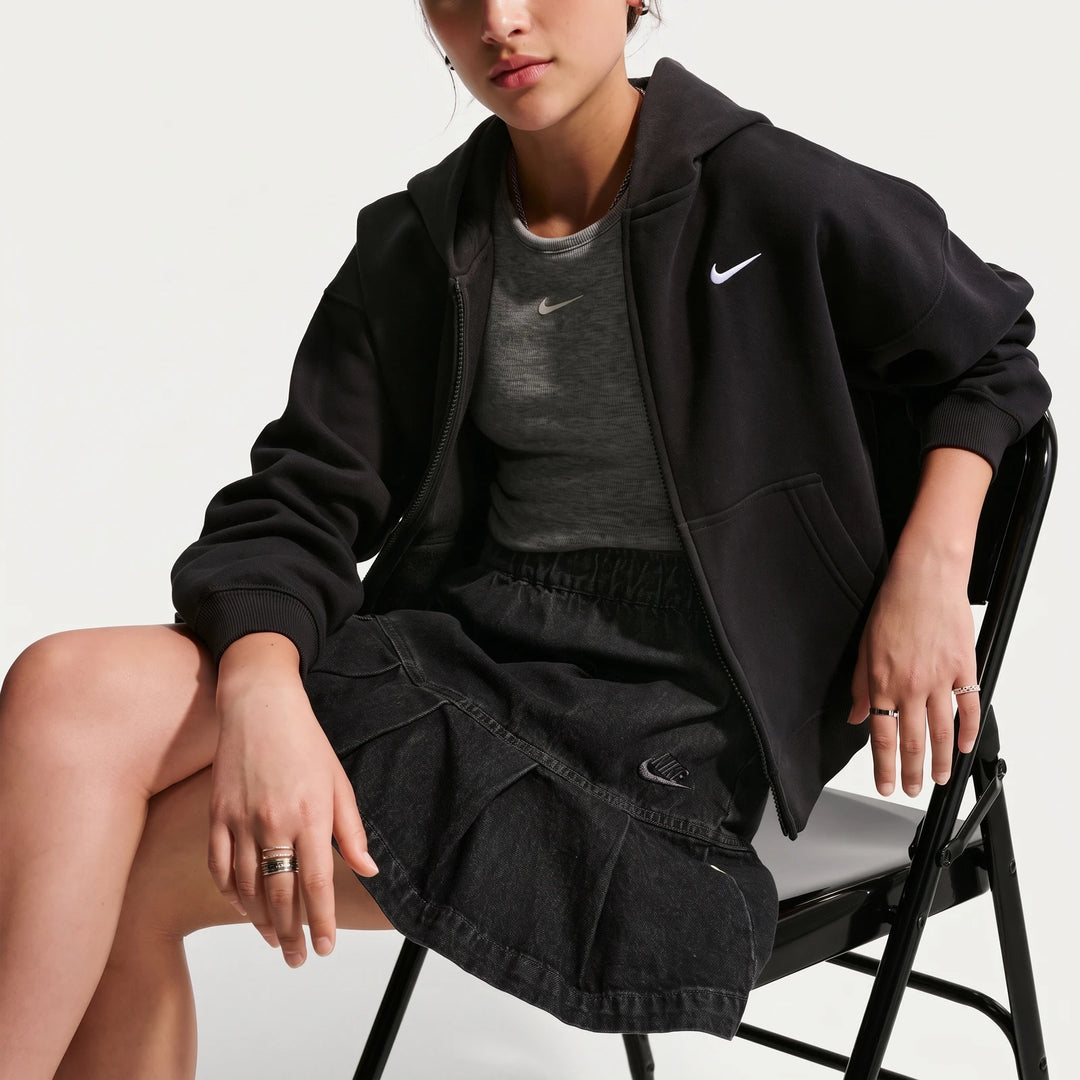 Nike Sportswear Big Kids' black denim pleated skirt worn by a girl, seated, also wearing a black full-zip hoodie and a grey