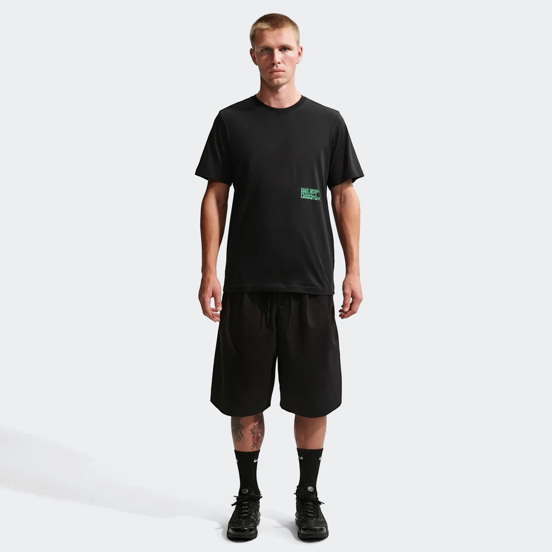 A male model wears a Nike Sportswear black t-shirt with a green graphic, black shorts, and black sneakers.