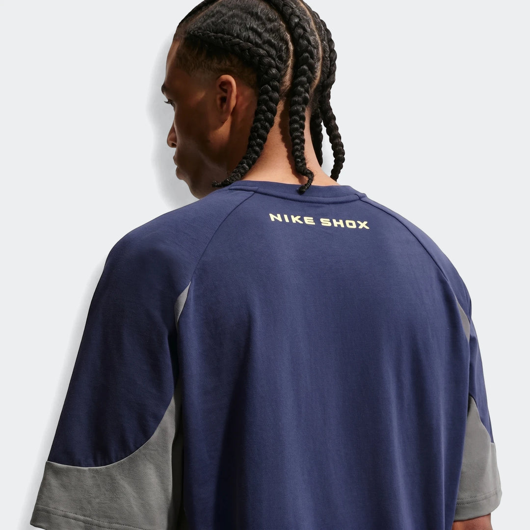 Rear view of a man wearing a navy blue Nike Sportswear short-sleeve top with gray panels and a yellow Nike Shox logo.