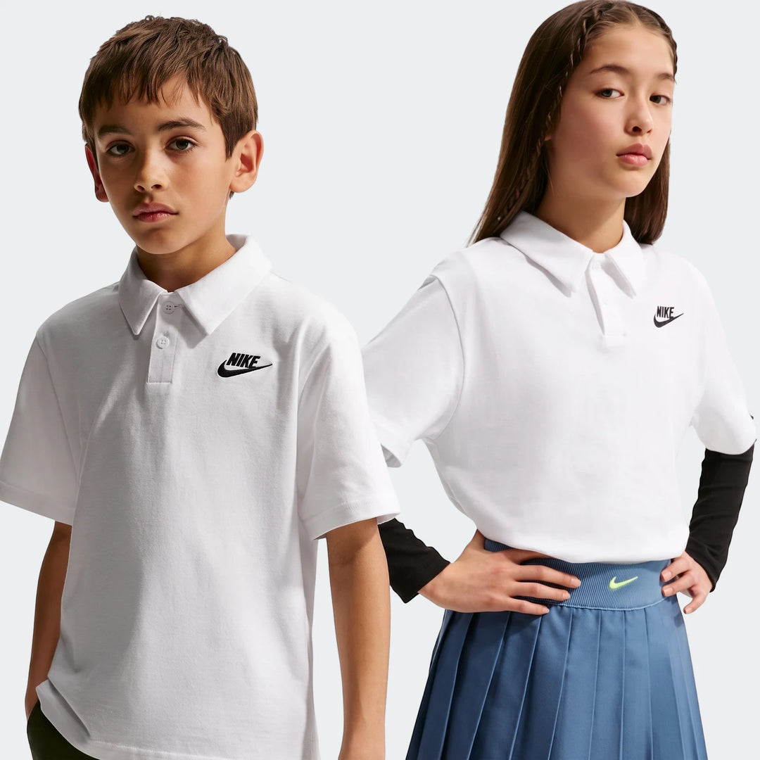 Two kids model white Nike Sportswear Club Big Kids' Knit Short-Sleeve Polo shirts; a boy in shorts and a girl in a pleated