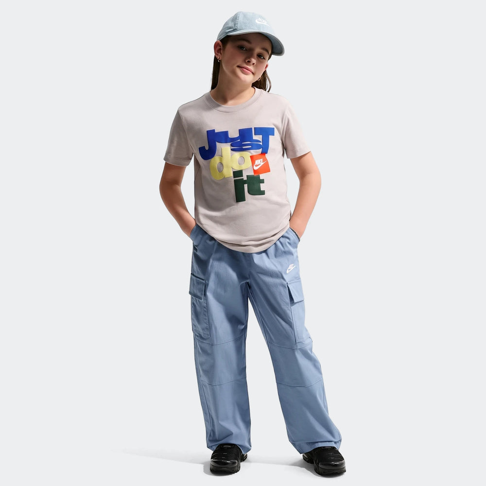 Young girl wearing a gray Nike Sportswear Club Big Kids' T-Shirt with a colorful 'Just Do It' graphic, blue cargo pants, and