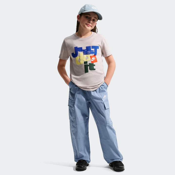 Young girl wearing a gray Nike Sportswear Club Big Kids' T-Shirt with a colorful 'Just Do It' graphic, blue cargo pants, and