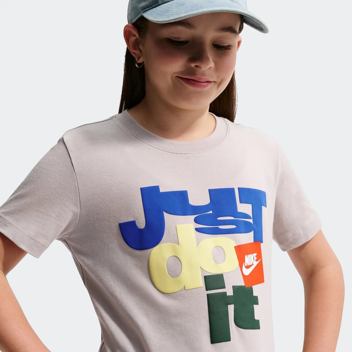 Young girl wearing a light grey Nike Sportswear Club Big Kids' T-Shirt with a colorful Just Do It graphic, accessorized with