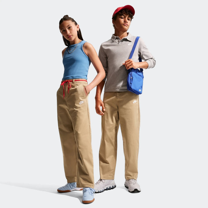 Two kids wearing Nike Sportswear Club Big Kids' Woven Everyday Pants in khaki with Nike logos, paired with casual tops and