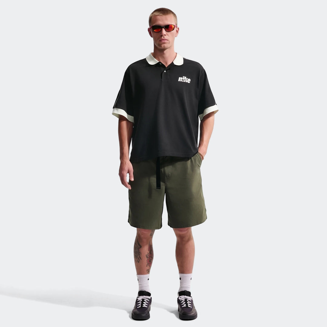 Full-body shot of a male model wearing a black Nike Sportswear Club oversized short-sleeve polo shirt, olive green shorts,