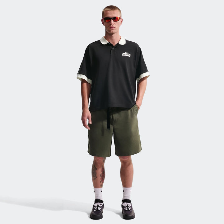 Full-body shot of a male model wearing a black Nike Sportswear Club oversized short-sleeve polo shirt, olive green shorts,