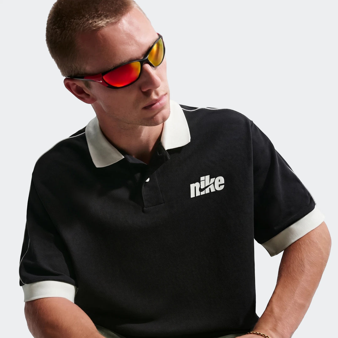 A man wears a Nike Sportswear Club black oversized short-sleeve polo shirt with white trim and red mirrored sunglasses,